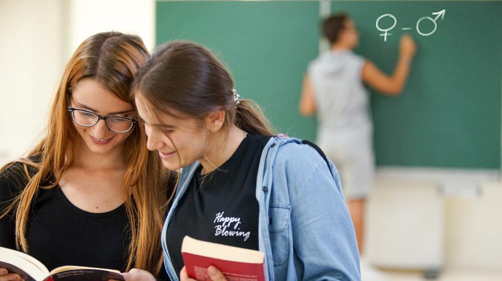 Two girls reading a book in the classroom, while a boy behind them is drawing the symbols female, male on the black board.