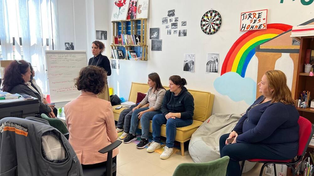 Several mothers sit in a room of the "Stay Together" community center in Kombinat, listening to the trainer explaining positive parenting information written on a board in front of her.