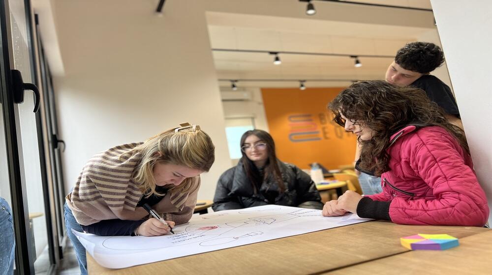 New generation of peacebuilders in Albania during a training. There are 3 girls sitting around a table, one of them is writting a poster. Also a boy is standing around the table watching the girl writting.