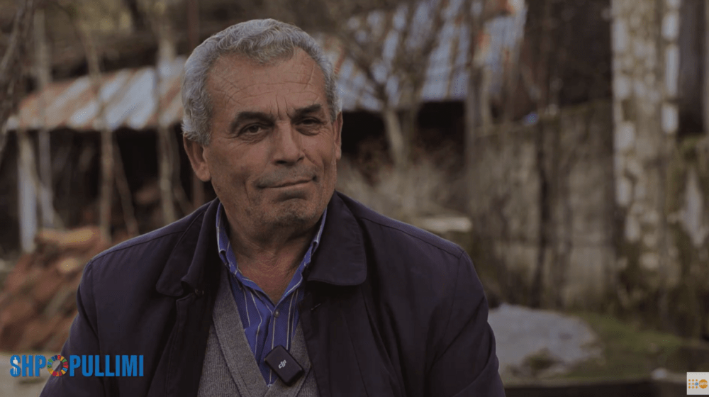 An elderly man being interviewed, in the background there is a view of the yard of his house surrounded by trees