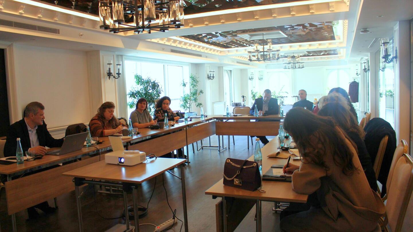 Meeting room with people around a U-shaped table, projector in foreground.